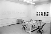 A photograph of a room with works on paper hung on one wall and photographs on the other wall. In the center of the room is a table, strewn with papers, with three chairs.