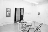 A photograph of a room with photographs and works on paper on the walls. In the center of the room is a table with folders and binders and three chairs. A door marked with 