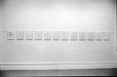 A photograph of a white wall hung with 12 works of the same size in a row.