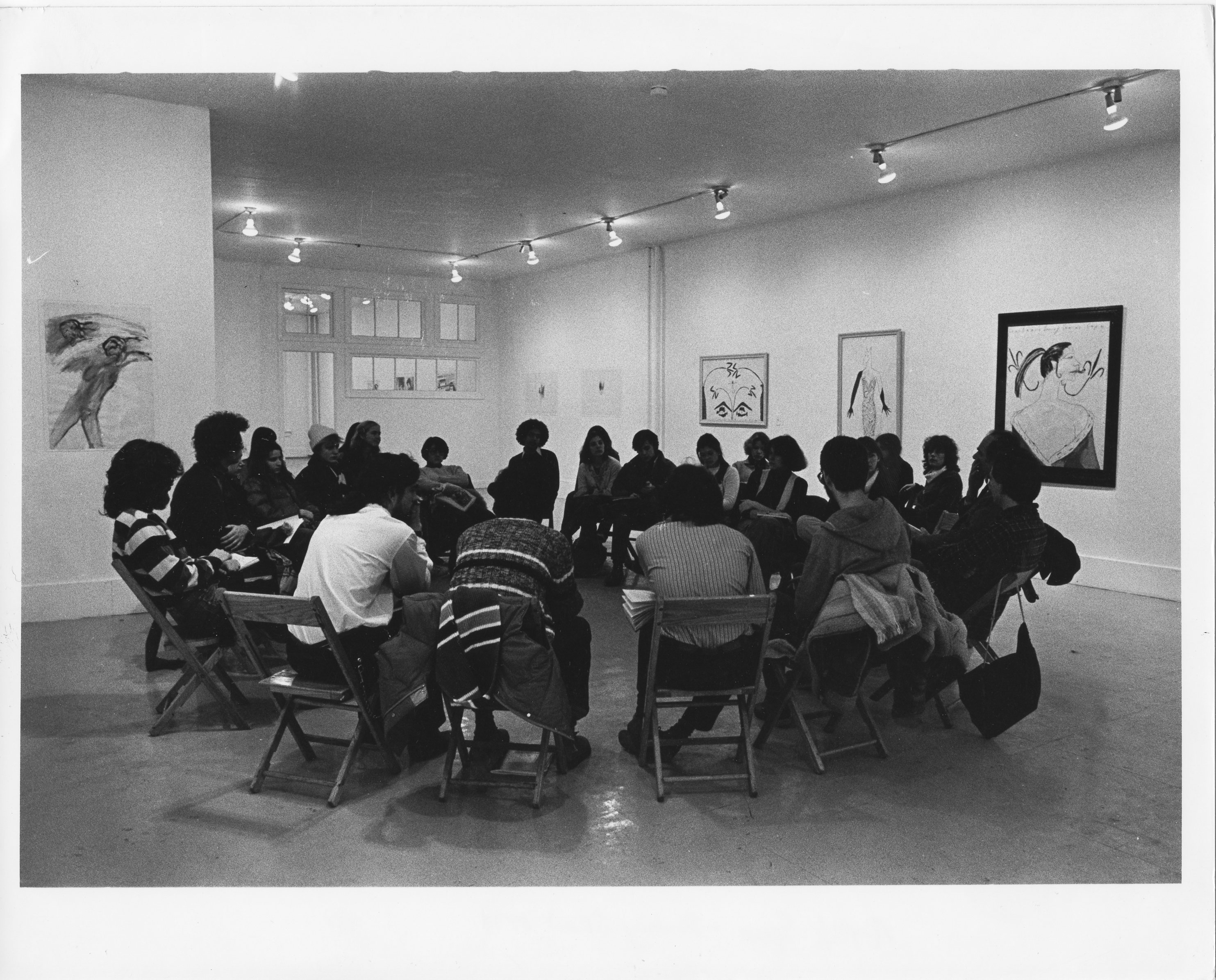 A group of people sitting in chairs arranged in a circle, all facing inwards. Drawings are hung on the walls.