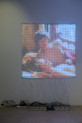Color image of an art work made up of 7656 small LED lights placed on a bleached fabric, creating an image of two figures laying intimately in bed together. Along the floor below the image are a string of wires, attached to various outlets and a controller.