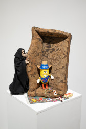 Color image of a sculpture sitting on a white pedestal. A beige couch is placed on its side, with a cowboy twinkie figurine sitting in the corner of the couch. Behind the side of the couch stands a grim reaper figure peering over. Beneath the couch is a small rug, with various items (play cards, popcorn, etc.) strewn across it.