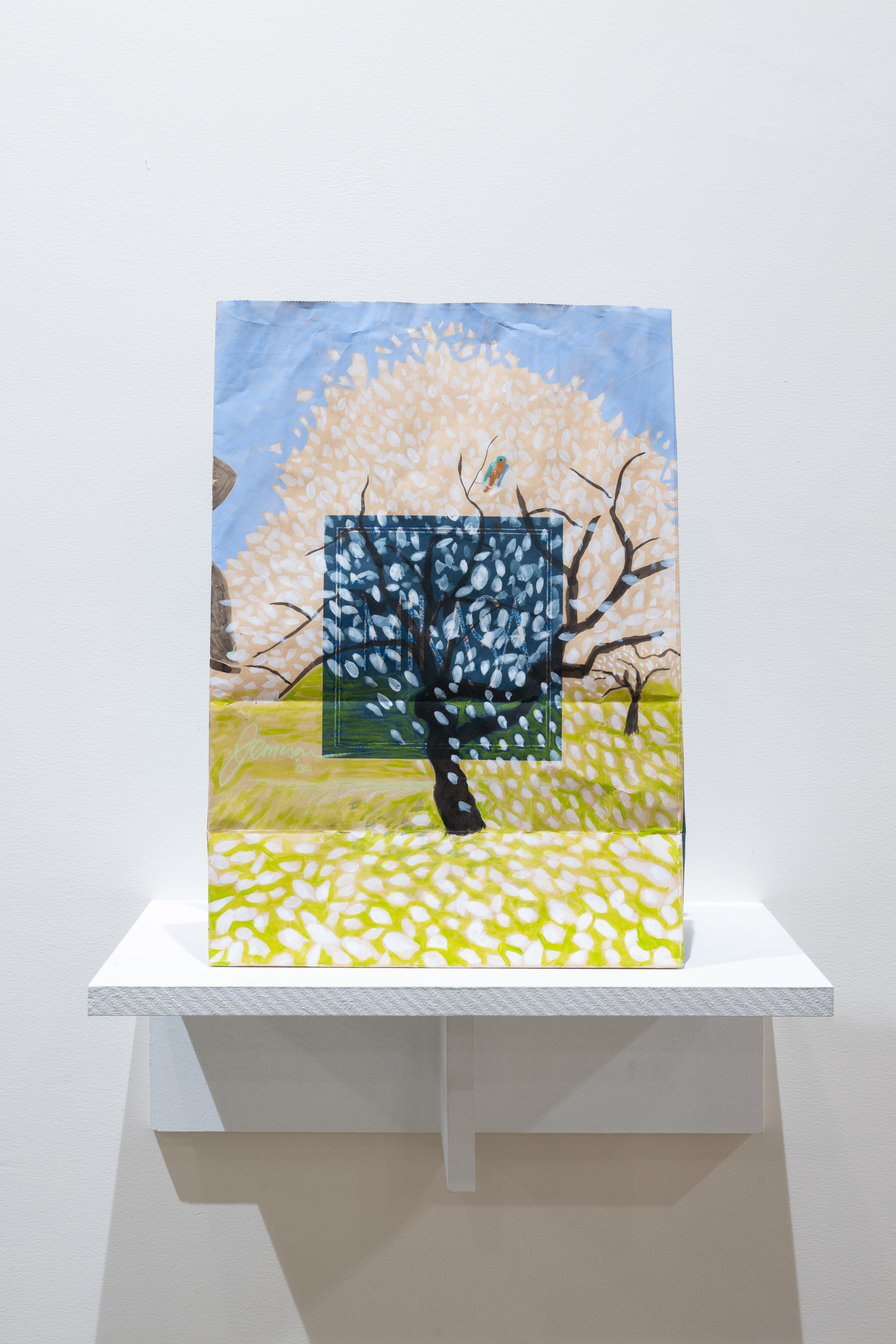 Color image of a painting, done on a paper bag, placed on a white pedestal. The painting features an apple blossom tree, with a light blue sky in the background, and green grass. The tree is painted over a blue square in the middle of the bag that reads 
