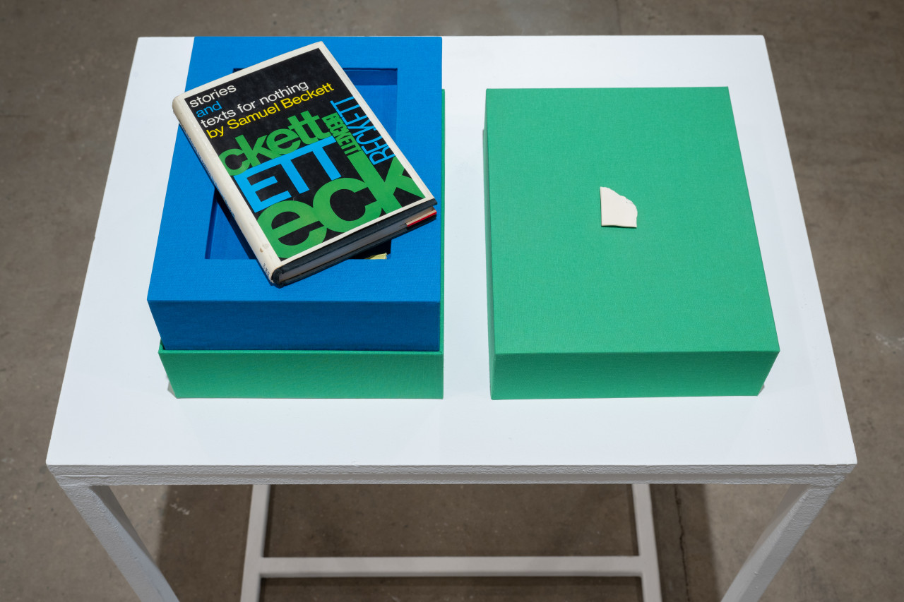 Color image of a bright green cloth-lined box with a bright blue interior, placed on a white pedestal. On one side of the opened box lies a copy of "stories and texts for nothing" by Samuel Beckett. On the top side of the box is a small piece of torn paper.