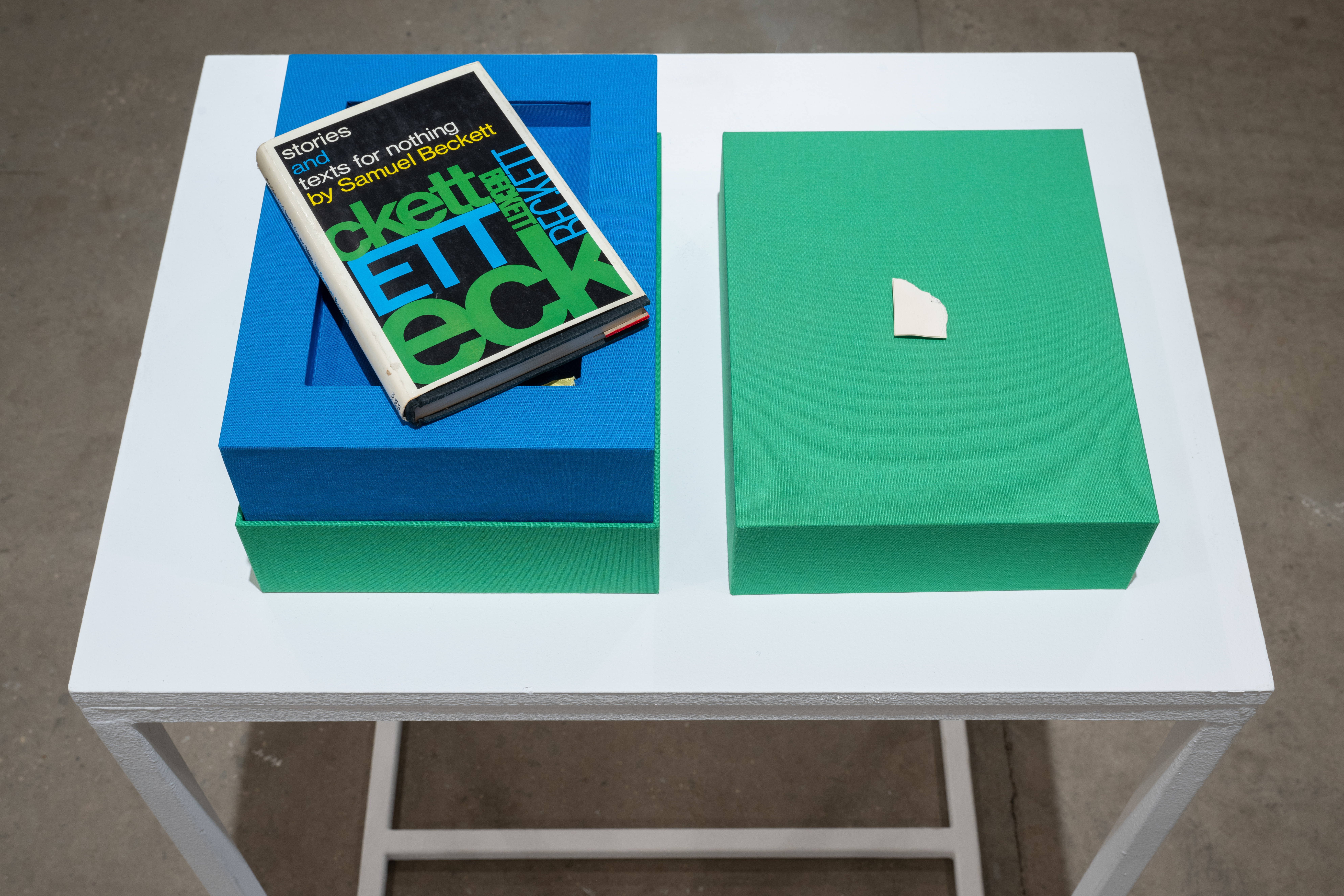 Color image of a bright green cloth-lined box with a bright blue interior, placed on a white pedestal. On one side of the opened box lies a copy of 