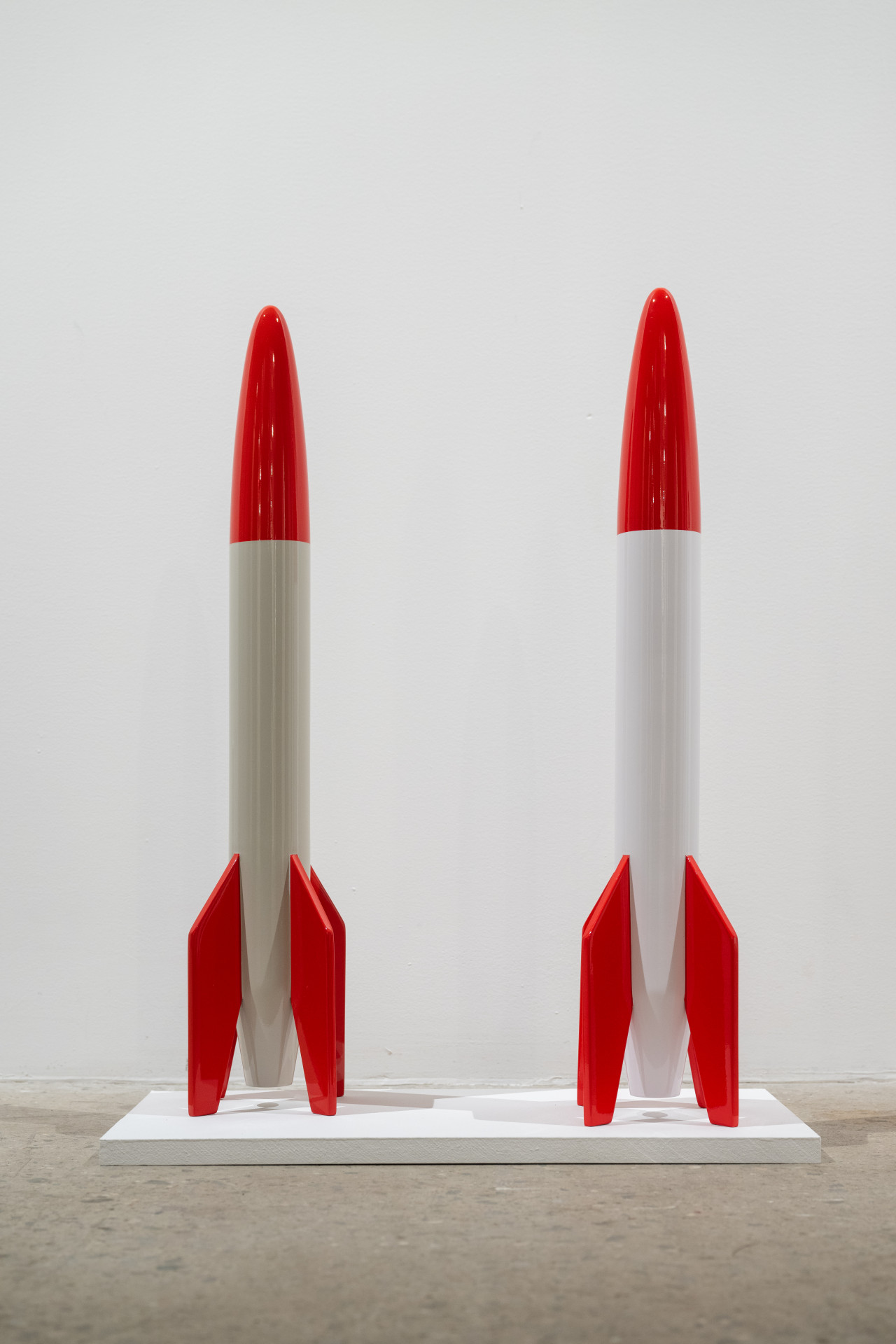 Color image of two replica rockets, created from wood and white and red enamel paint over top. They are placed on a small white platform.