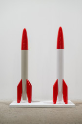 Color image of two replica rockets, created from wood and white and red enamel paint over top. They are placed on a small white platform.