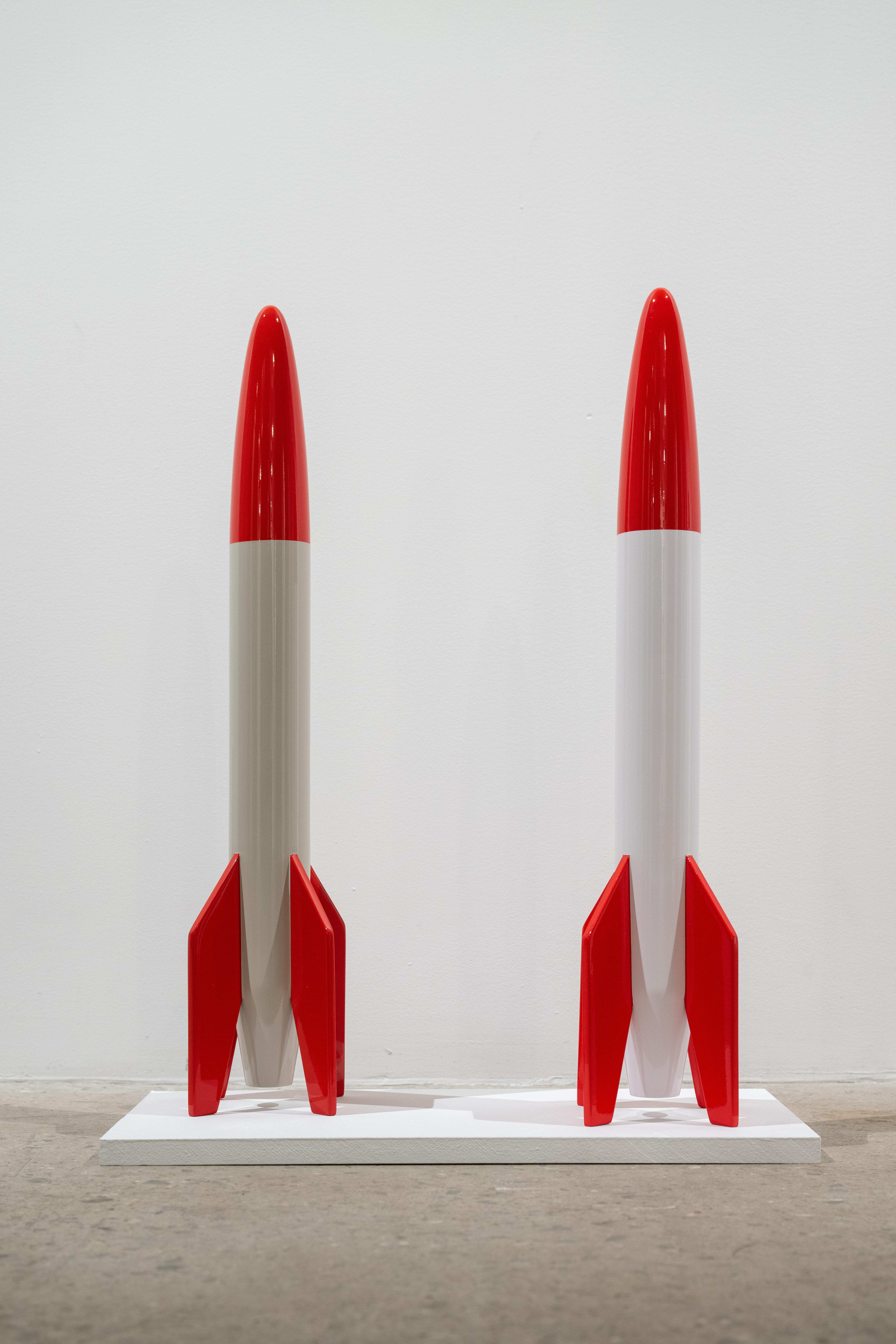 Color image of two replica rockets, created from wood and white and red enamel paint over top. They are placed on a small white platform.