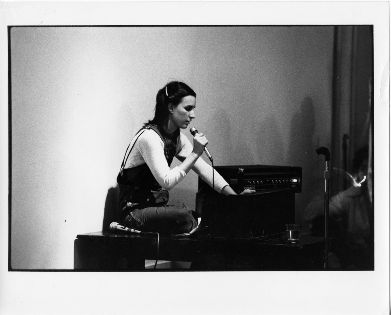 A black and white photograph of a figure sitting on a table. They hold a microphone in their right hand, and extend their left hand down behind a dark object on the surface of the table.