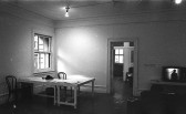 A black and white photograph of a gallery space. On the left, a window casts soft light onto a table and two chairs. On the right, a television monitor is positioned on a low bench.