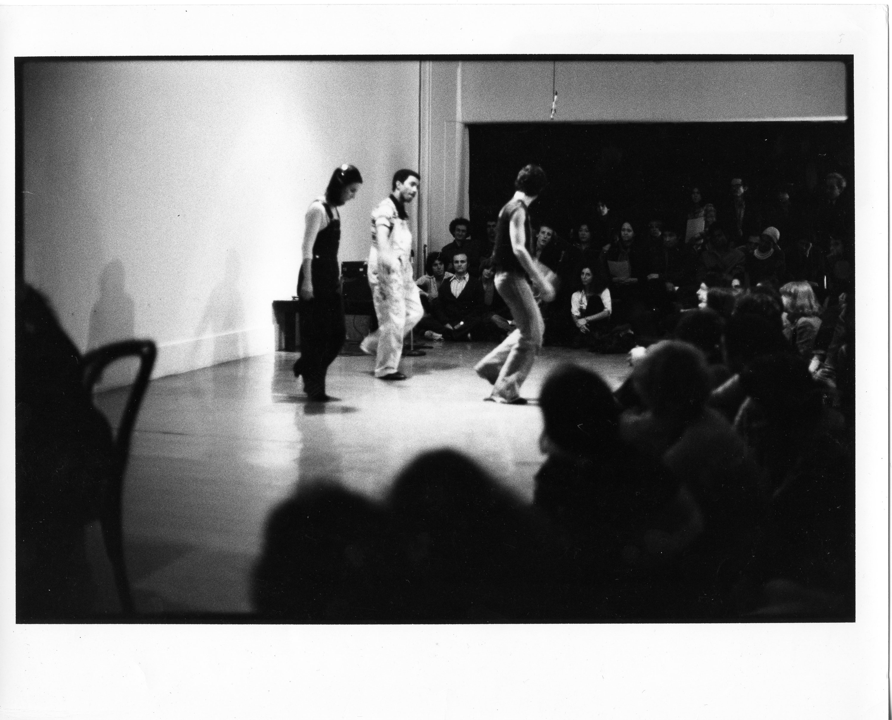 A black and white photograph of a performance. Three figures stand in a cleared portion of a room, each one balancing on their right foot. A crowd looks on.