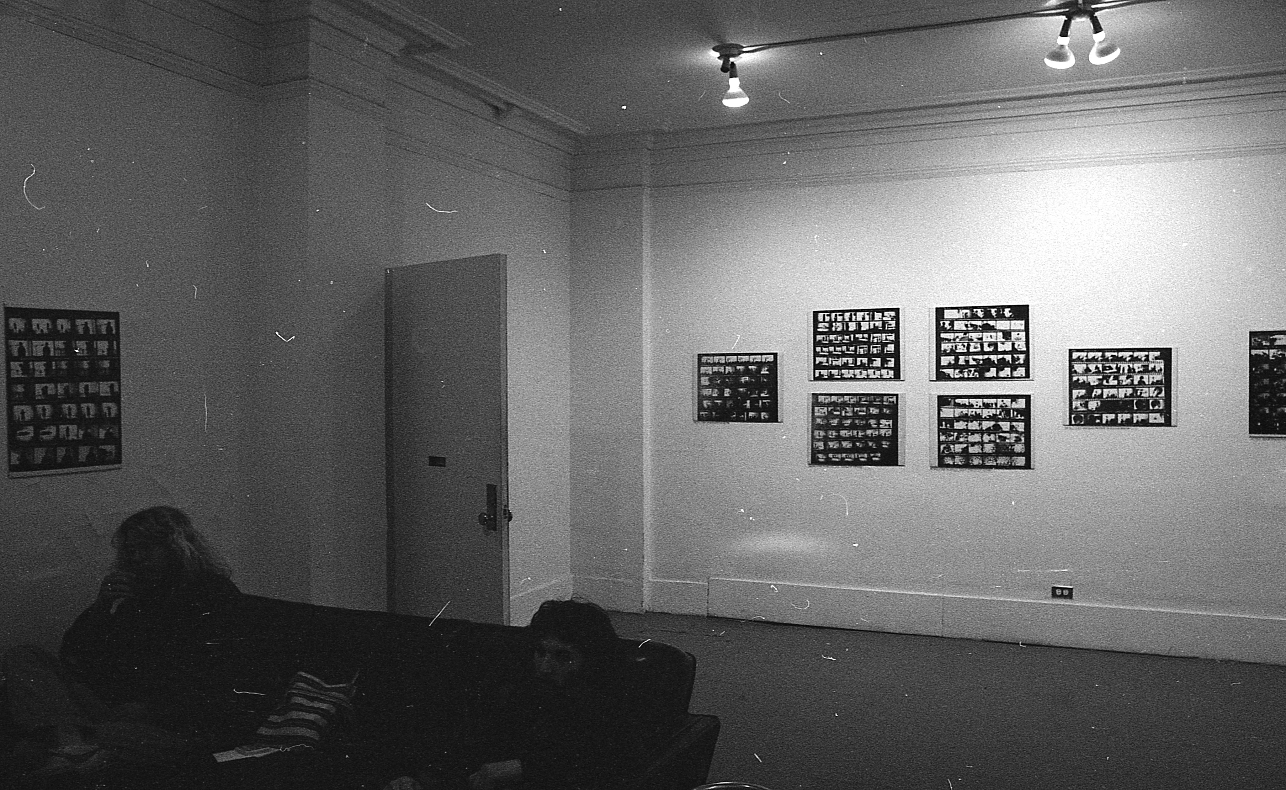 A black and white photograph of a gallery space. A chouch with two figures is visible in the lower left corner. On the walls, several framed works are hung, each with a grid of smaller black and white images.