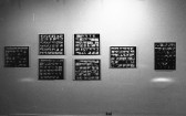 A black and white photograph of a gallery space. On the wall, seven framed works are hung, each featuring a grid of small black and white images.