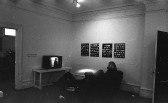 A black and white photograph of a gallery space. A television monitor is placed on a bench on the left, and two figures look on from a couch directly across from the screen. Hanging on the wall to their right are four framed works.