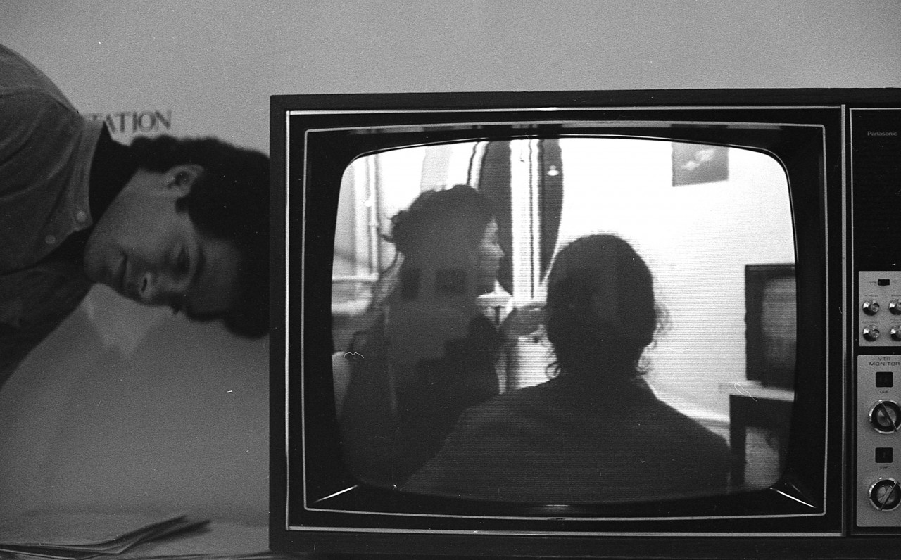 A bends at the waist, resting their head on the side of a television monitor. The monitor