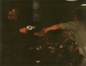 A color photograph of two figures in a dark space. The figure on the left wears a mask and points a gun at the figure on the right who is behind a bar.