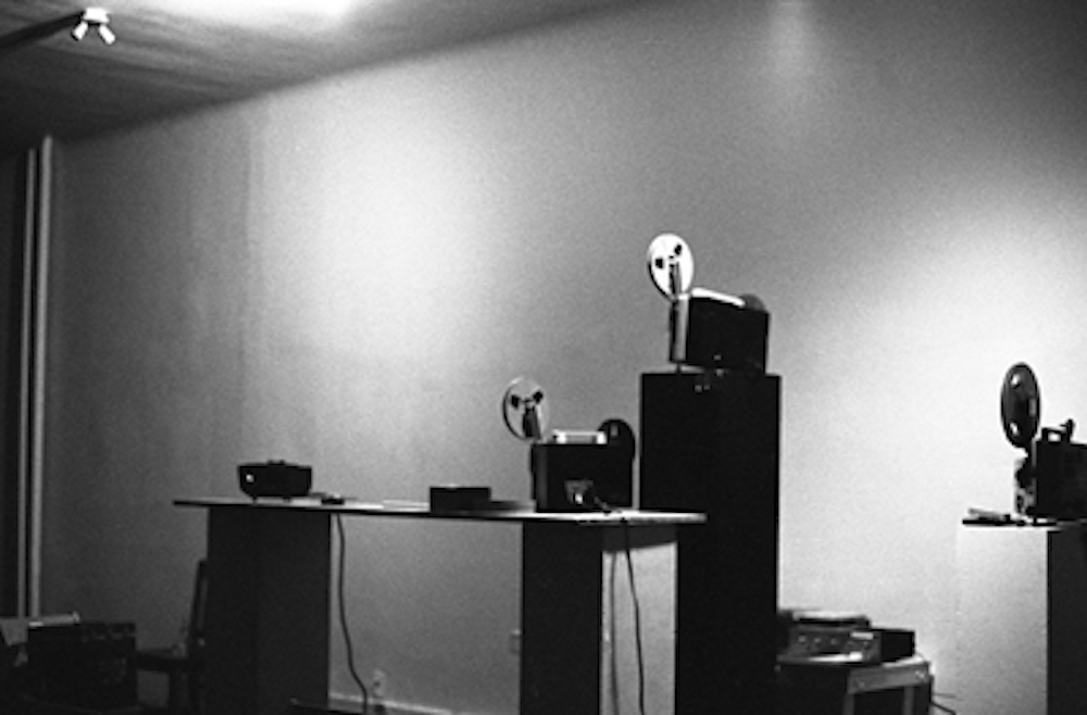 A black and white photograph of a gallery space. Several film reels and projectors are positioned on tables and stands.