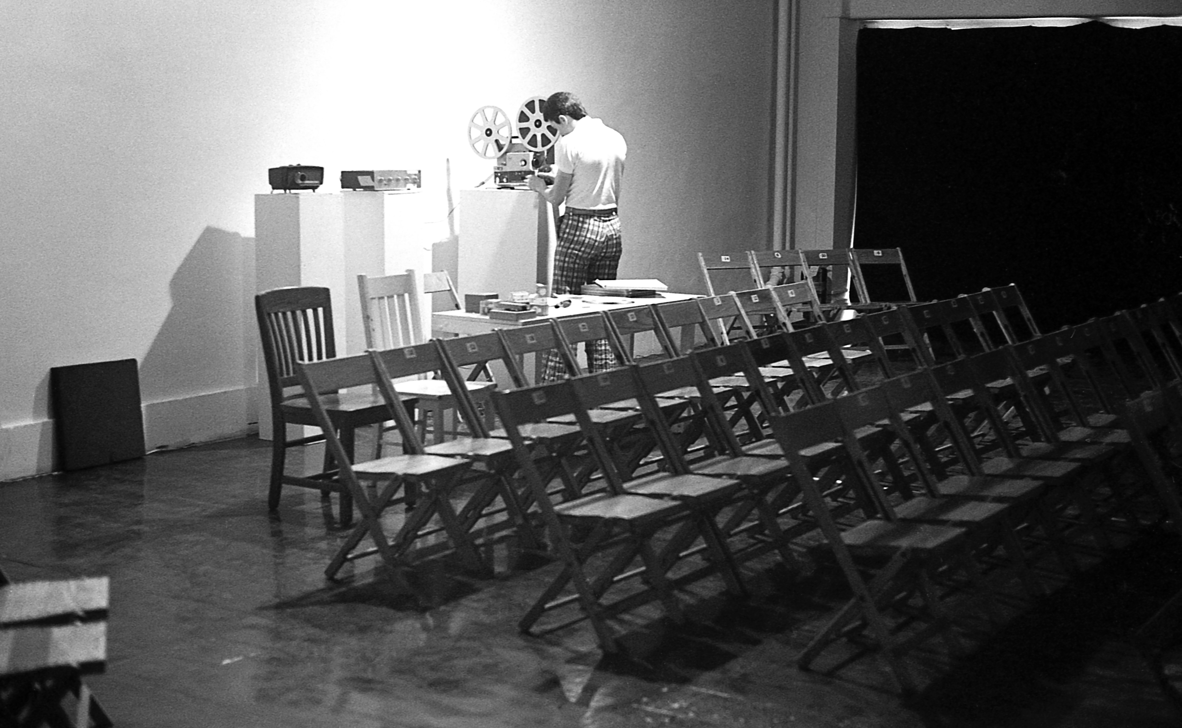 A black and white photograph of a room with several rows of chairs. Along the back wall, two projectors stand on pedastals and a figure on the right adjusts a film reel.