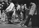 A black and white video still of a group of people in a street. One is sitting on a bicycle, another to the right leans against a trash can and looks down at their shoes.