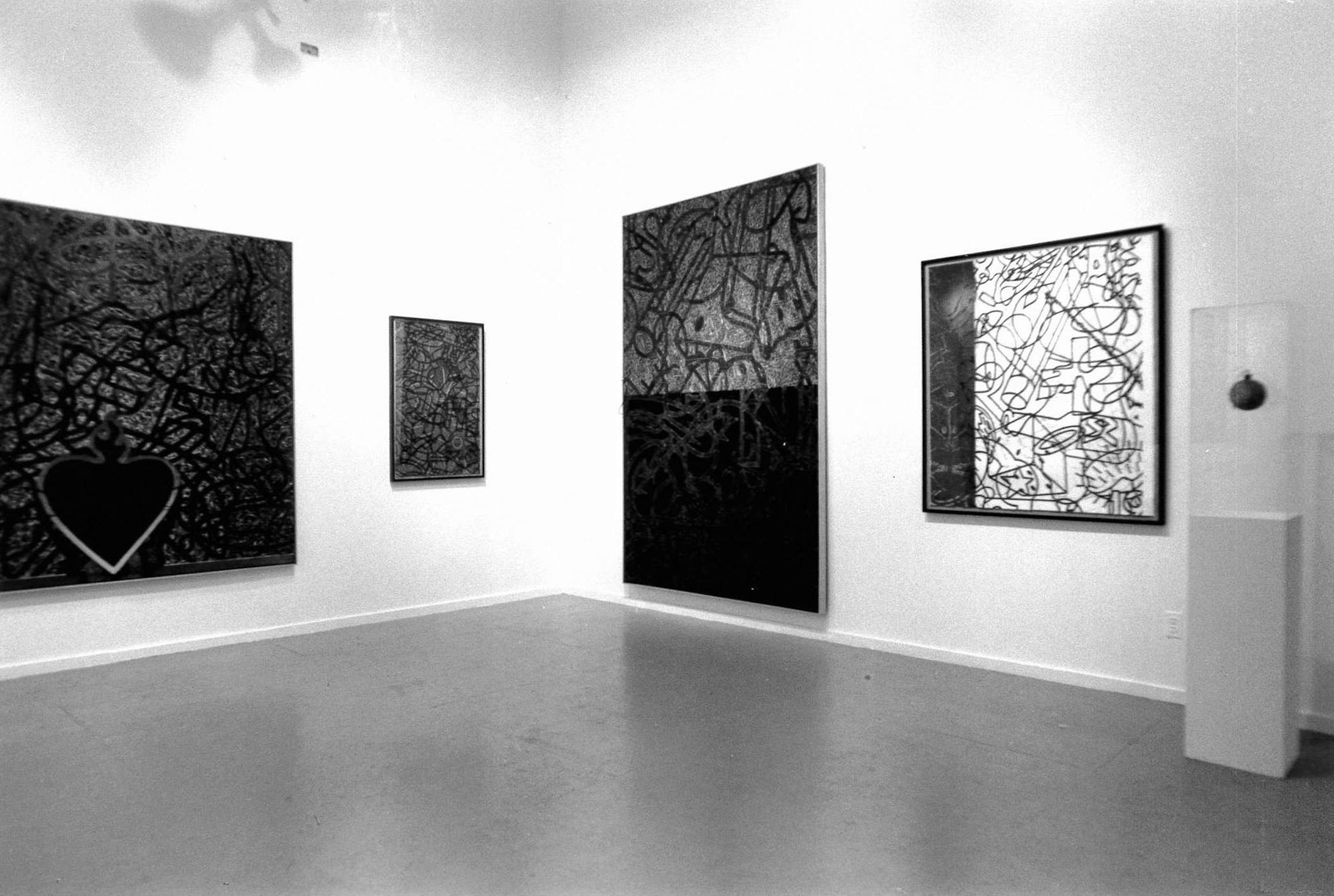 A view of a white-walled gallery with large abstract paintings hanging on the walls and a small pedestal display case on the right.