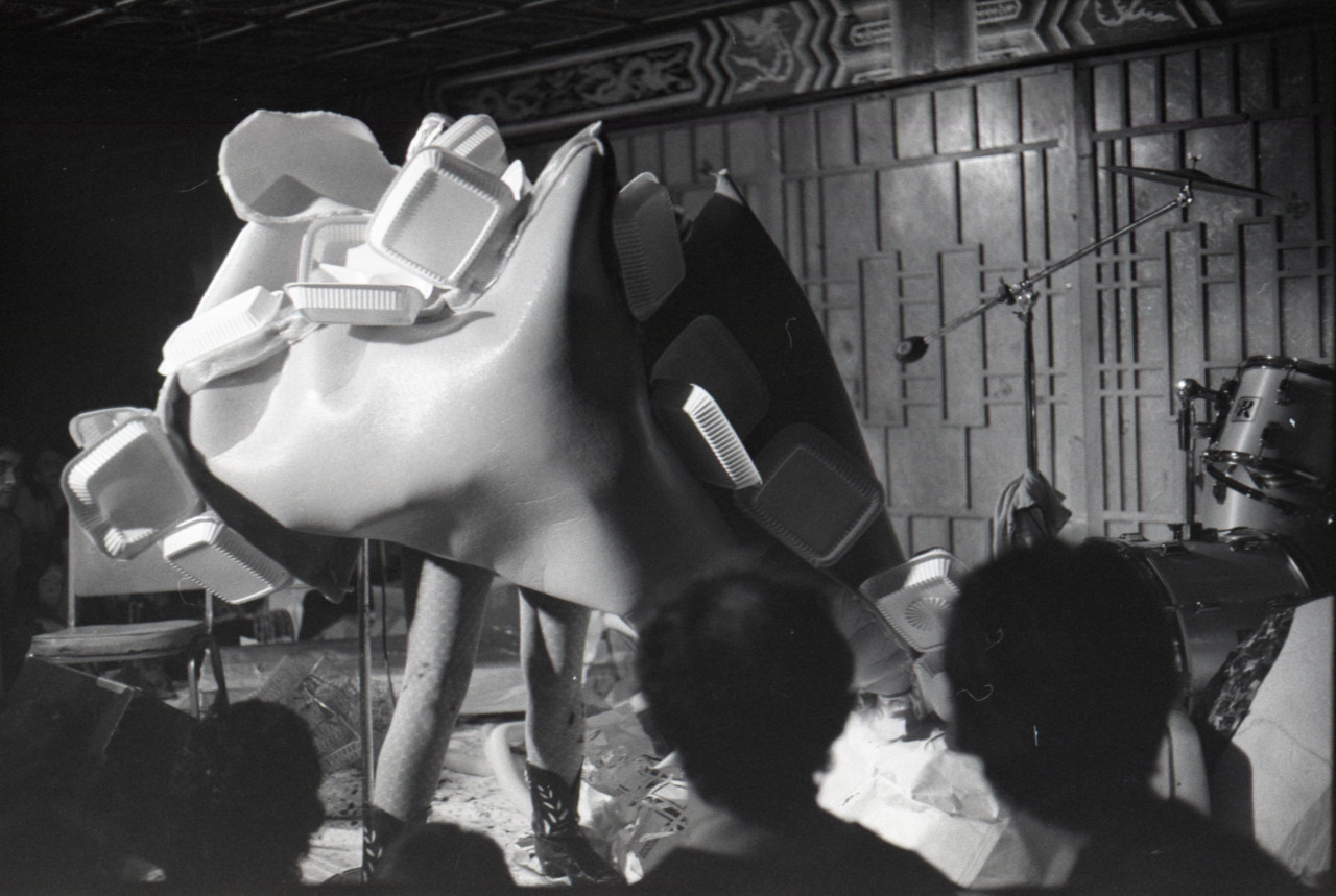 Black and white image depicting a performance. The artist is standing in the center of the frame and wearing spotted tights and cowboy boots. The upper half of their body is enveloped in a large plastic foam fabric, which covers their torso, face, and arms. Affixed to the large fabric construction are clusters of plastic takeout containers. Audience members are visible in the foreground.