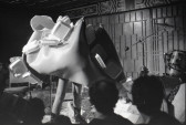Black and white image depicting a performance. The artist is standing in the center of the frame and wearing spotted tights and cowboy boots. The upper half of their body is enveloped in a large plastic foam fabric, which covers their torso, face, and arms. Affixed to the large fabric construction are clusters of plastic takeout containers. Audience members are visible in the foreground.