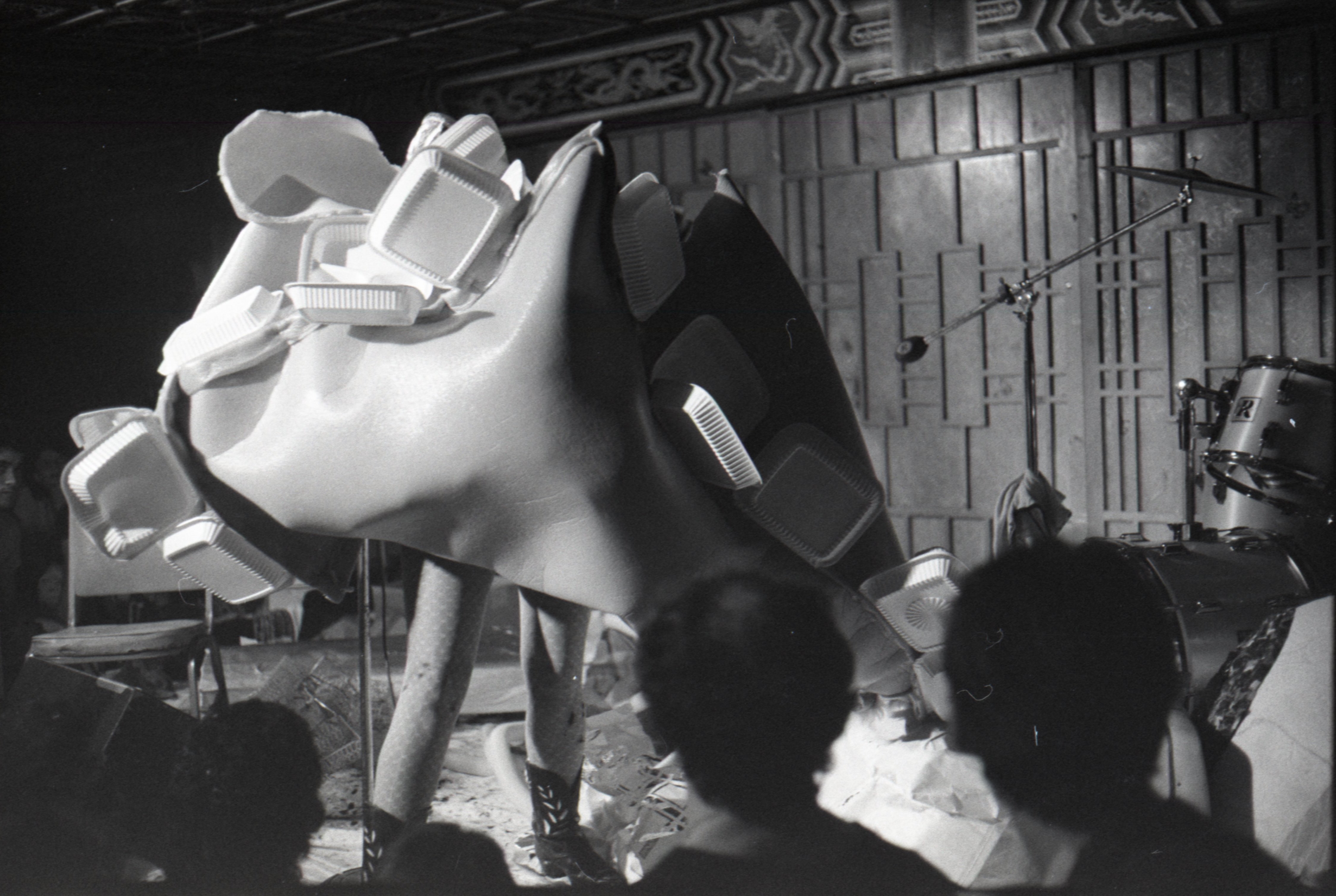 Black and white image depicting a performance. The artist is standing in the center of the frame and wearing spotted tights and cowboy boots. The upper half of their body is enveloped in a large plastic foam fabric, which covers their torso, face, and arms. Affixed to the large fabric construction are clusters of plastic takeout containers. Audience members are visible in the foreground.