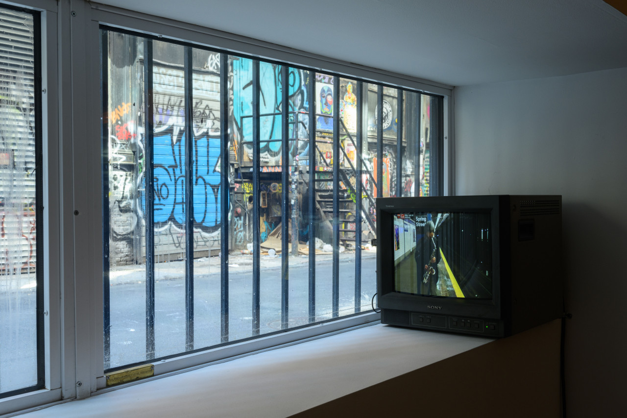 A small television set sits in front of a glass window with bars. Behind the window, grafitti can be seen.