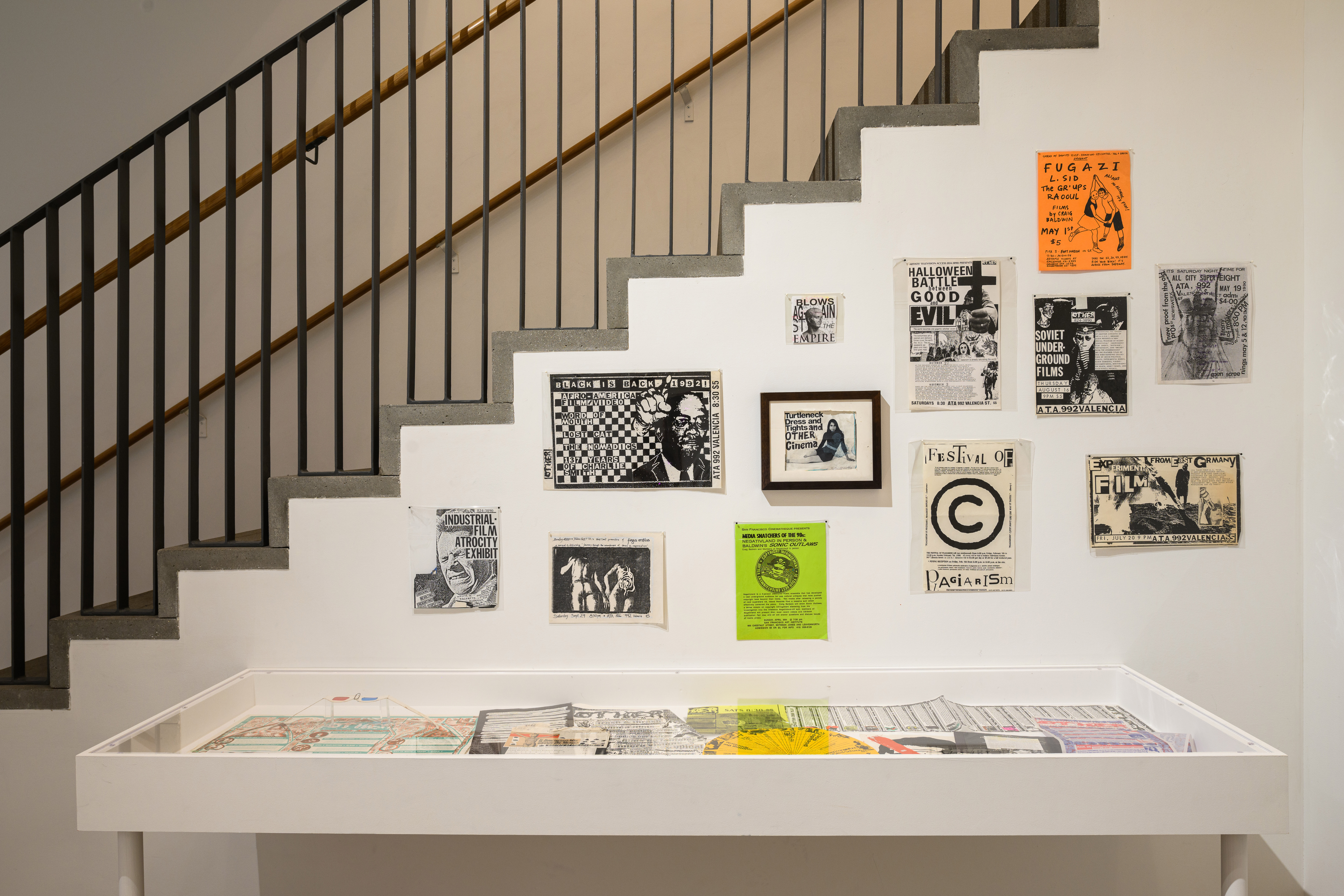 An image of a glass display case, above which hang various posters and epehemera. Behind the posters is a staircase.