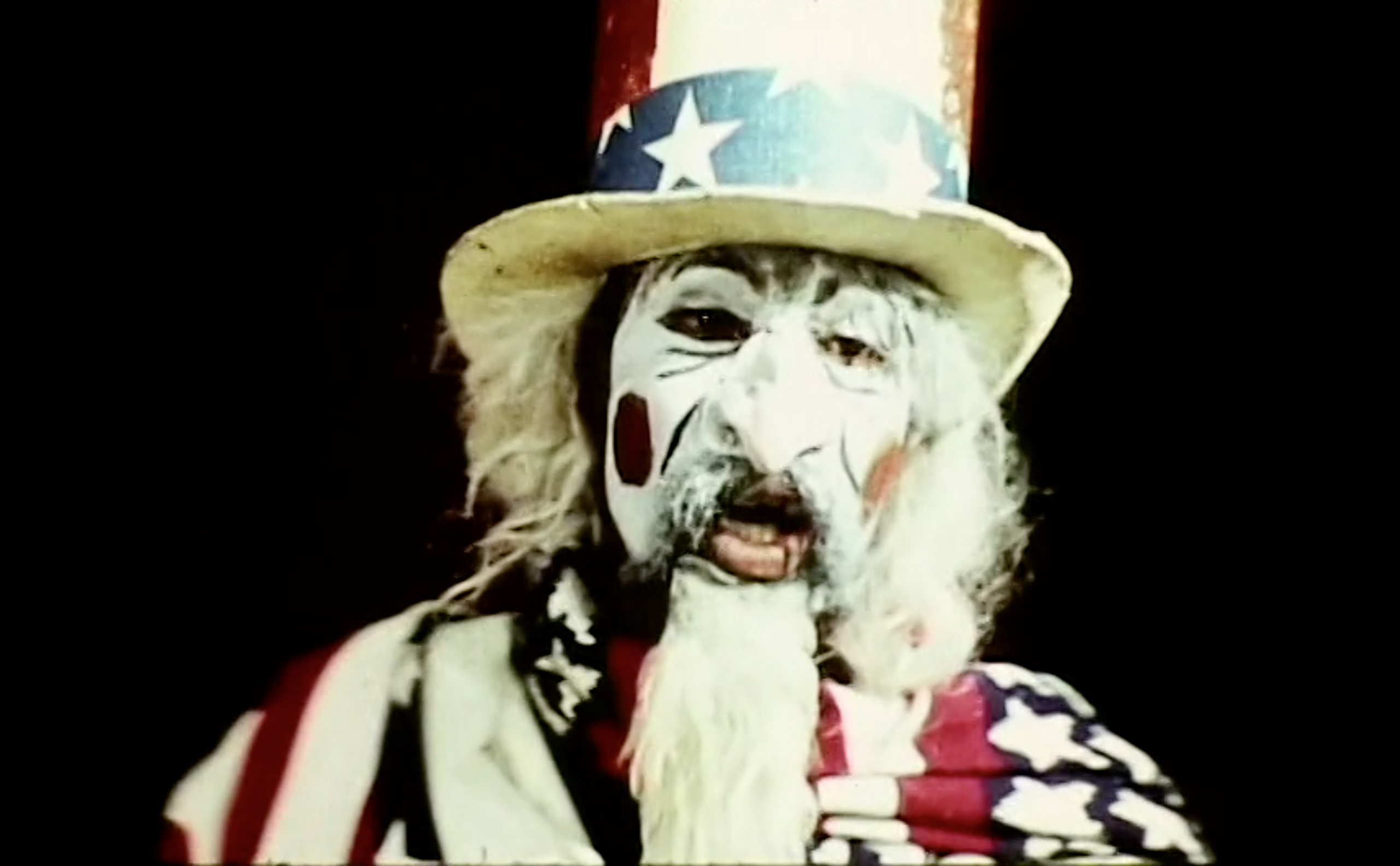A colored image depicts a figure's upper half of the body facing front against a black background. The figure's face is painted white with contrasting makeup. The figure is wearing a fake white nose, a white wig, and an American flag patterned hat and scarf around his shoulder.