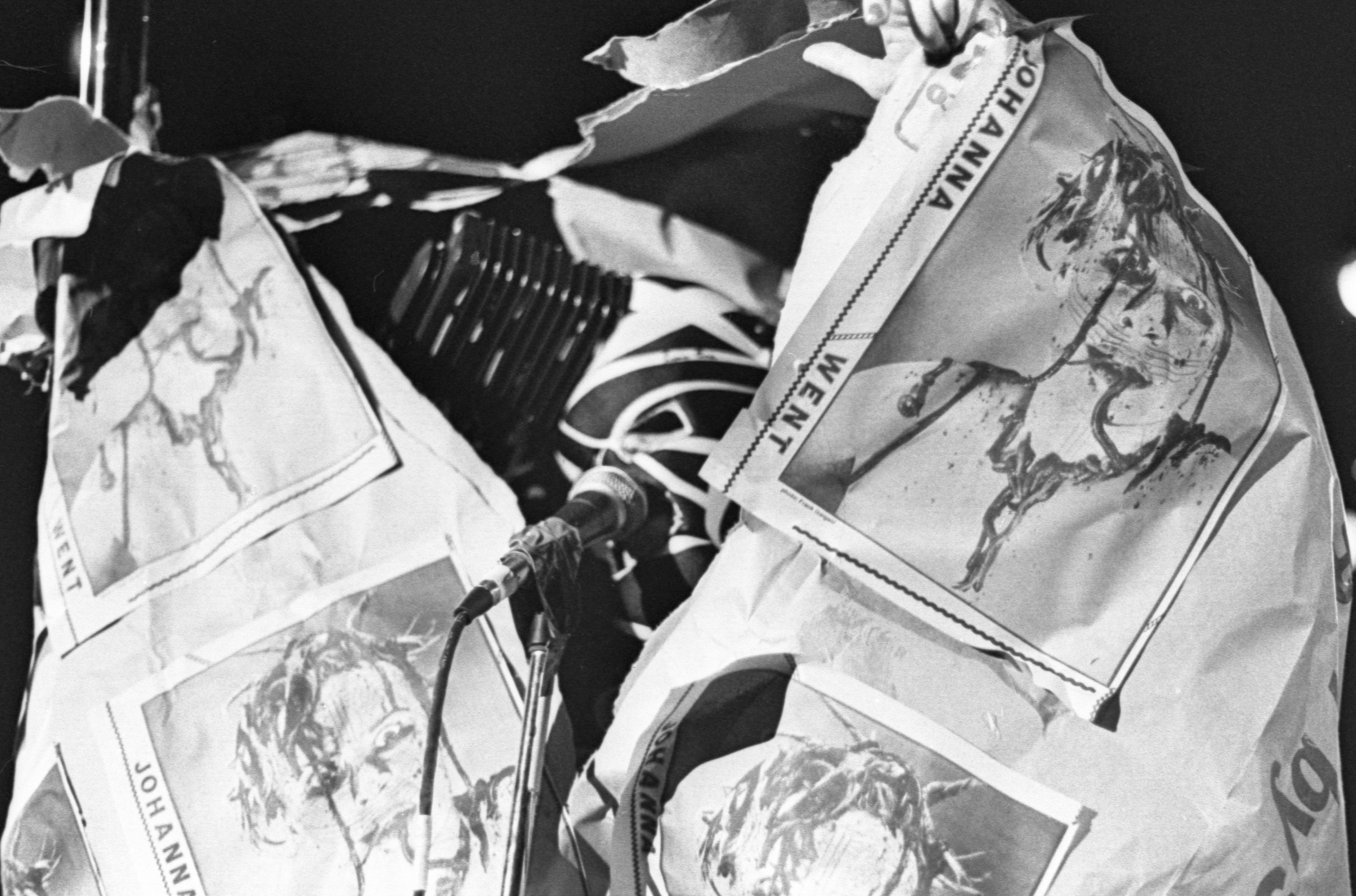 Black and white image of a performance. The artist is shown standing in front of a microphone, but their face is completely covered in a mask with criss-crossing white stripes. Their upper body is wrapped in a large sheet of thick paper with repeating images pasted onto its surface, and a square cone hat sits at an angle on their head.