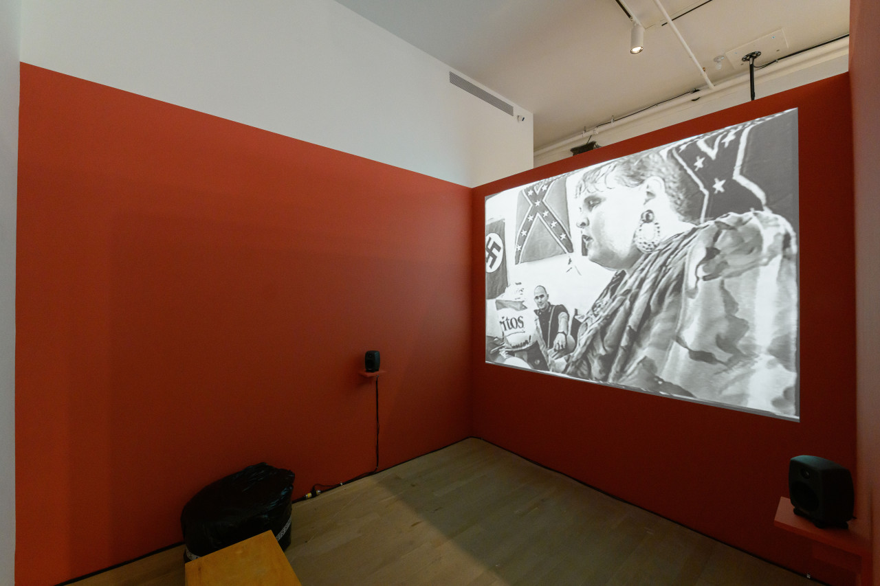 An image of a gallery space with walls painted red and a projection with speakers in front of the right-facing wall.