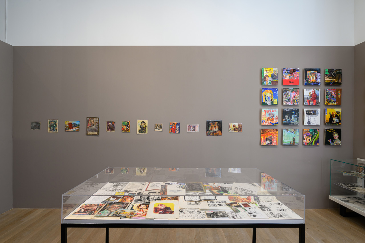 A gallery with a large glass display case in the center. On the wall behind it hang various ephemera.