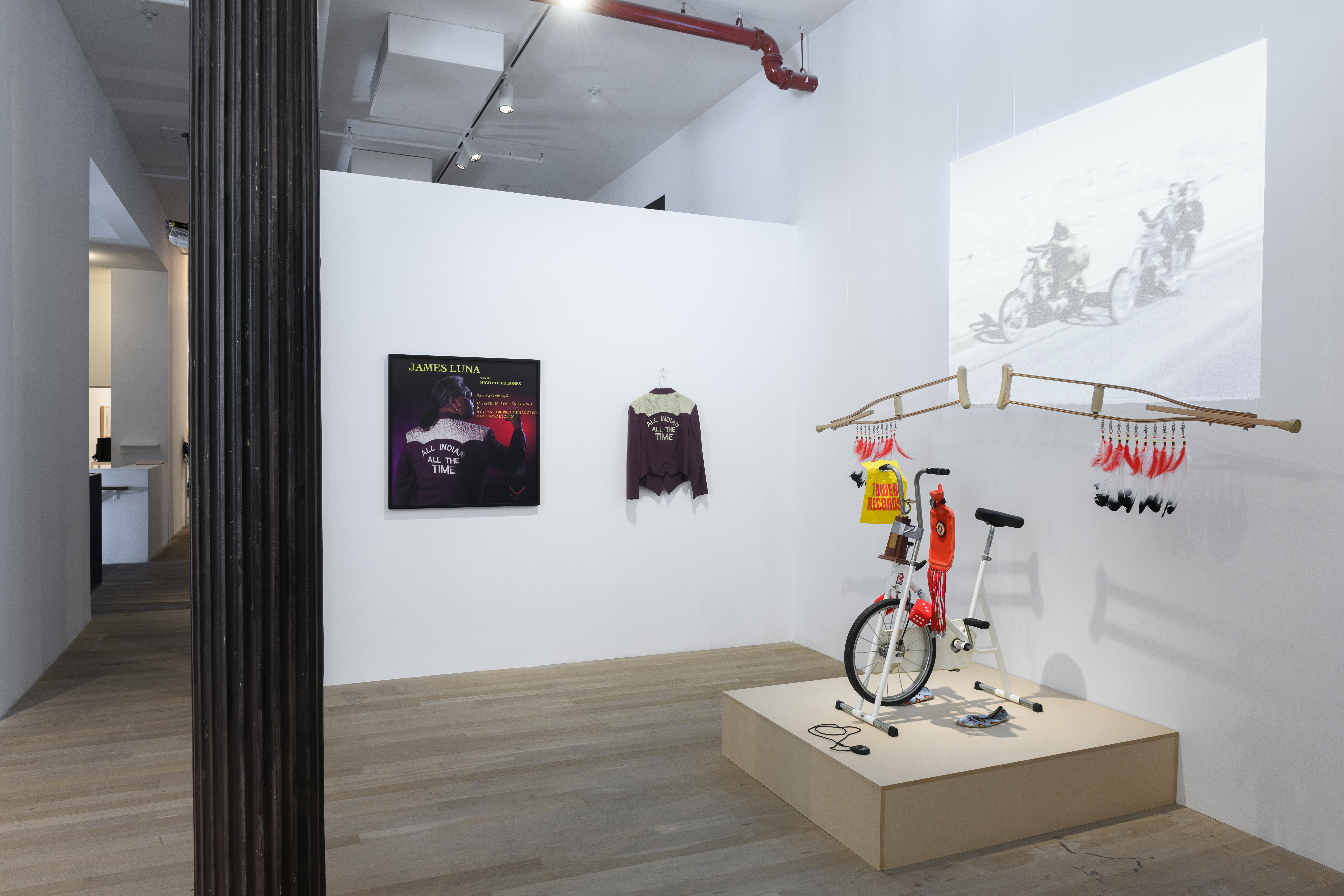 An image of a gallery with a jacket and poster hanging on one wall, and a projection visible on the other. In front of the projection is a sculpture made out of bike parts and other materials.