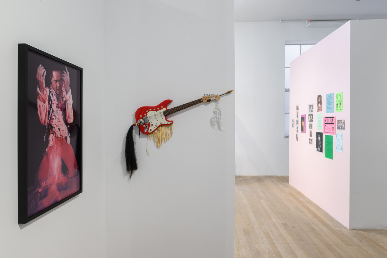 On the left, a framed photograph hangs on the wall. In the center, a sculpture built around a red electric guitar hangs from the wall. In the distance on the right, various posters and ephemera adorn the wall.