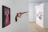 On the left, a framed photograph hangs on the wall. In the center, a sculpture built around a red electric guitar hangs from the wall. In the distance on the right, various posters and ephemera adorn the wall.