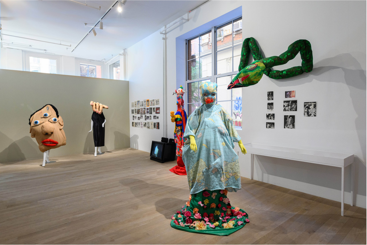 The walls on either side of a large glass window have various ephemera hanging from them. On the ground stand four large humanoid costumes, with a TV monitor behind them.