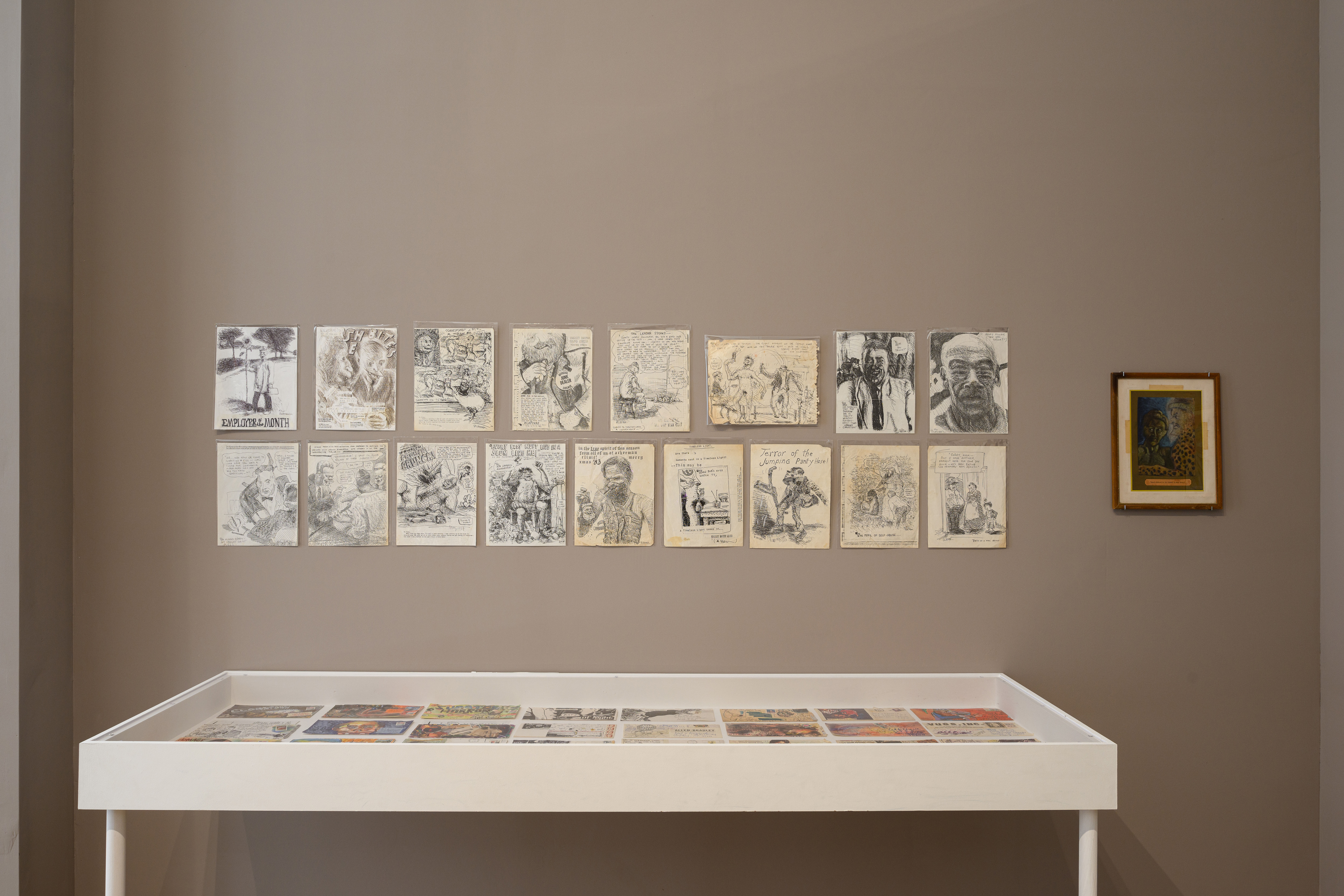 An image of a glass display case, above which several drawings hang from the wall.