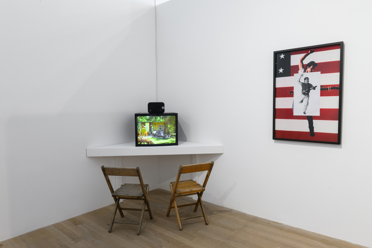 An image of a monitor with two chairs in front of it. To the right of the monitor, a photocollage hangs on the wall.