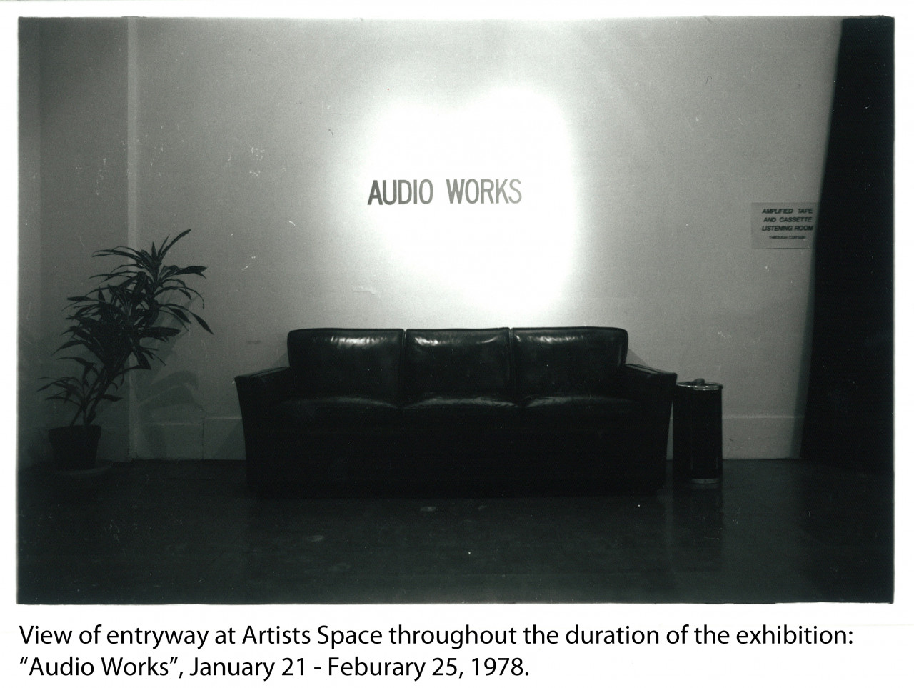 A dimly lit room with a black couch against the far wall and a houseplant in the left hand corner. Above the couch are the words 