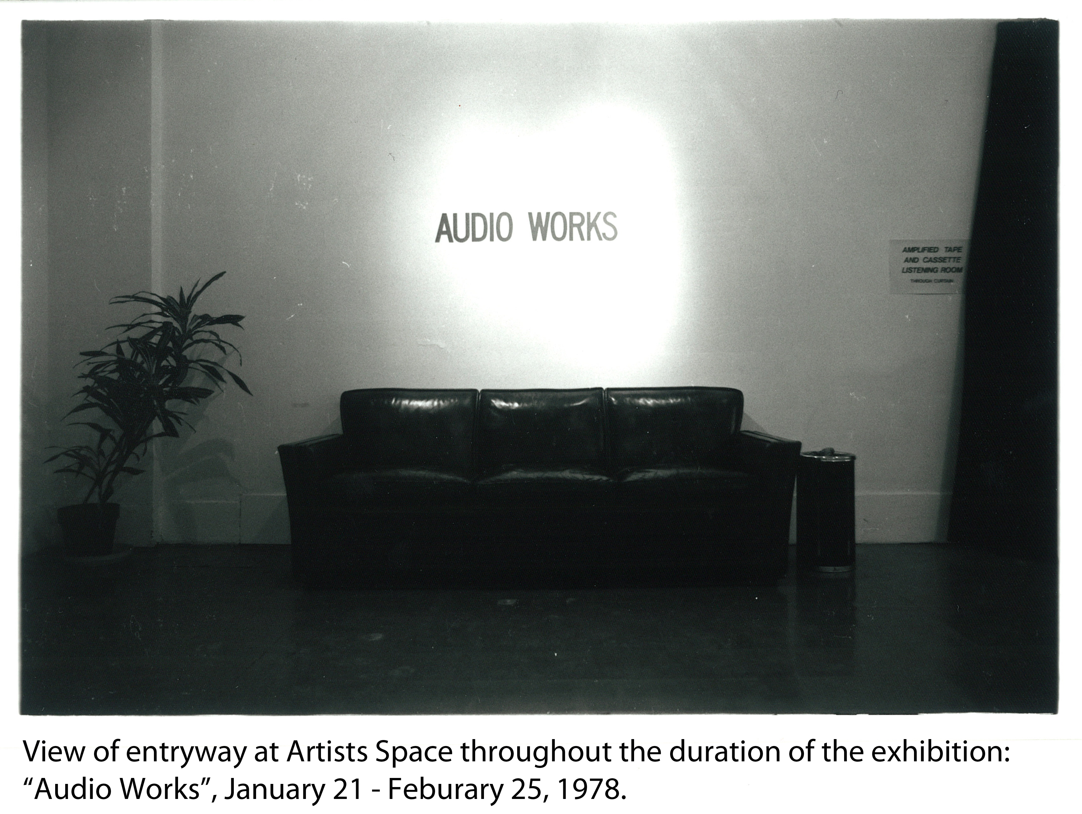 A dimly lit room with a black couch against the far wall and a houseplant in the left hand corner. Above the couch are the words 