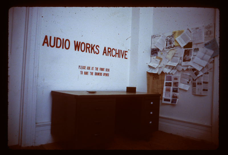 A room with a large set of drawers set against the wall. Above the drawers are the words "Audio Works Archive / Please Ask At The Front Desk To Have The Drawers Opened." On the right wall hang several papers and print-outs.