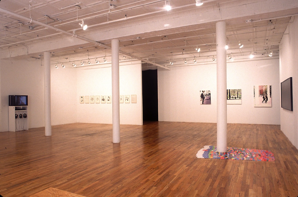 A wide shot of a gallery space with three columns. At the base of one column is a colorful, flat piece. On the large wall behind that column, three paintings hang in a horizontal line. To left of that, several framed drawings are arranged in a horizontal line. Against the perpendicular wall, there is a plinth with a television and several headphones.