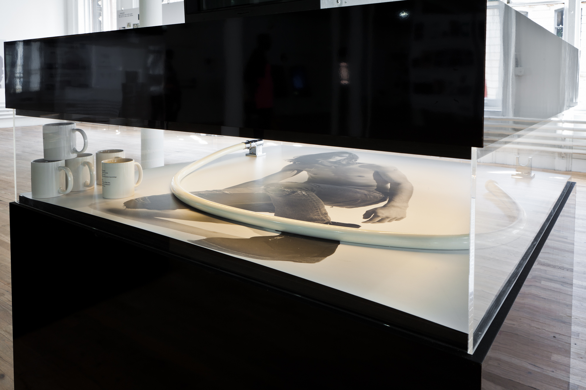 Detail view of the smaller display case, looking through a narrow glass window. Five mugs are stacked in a corner, and a short pipe curls around the center of the case. A black-and-white image of a shirtless man wearing ripped jeans and boots is printed on the surface.