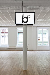 Two screens installed on either side of the top portion a white column in a gallery space. The front screen displays a logo consisting of a large letter 