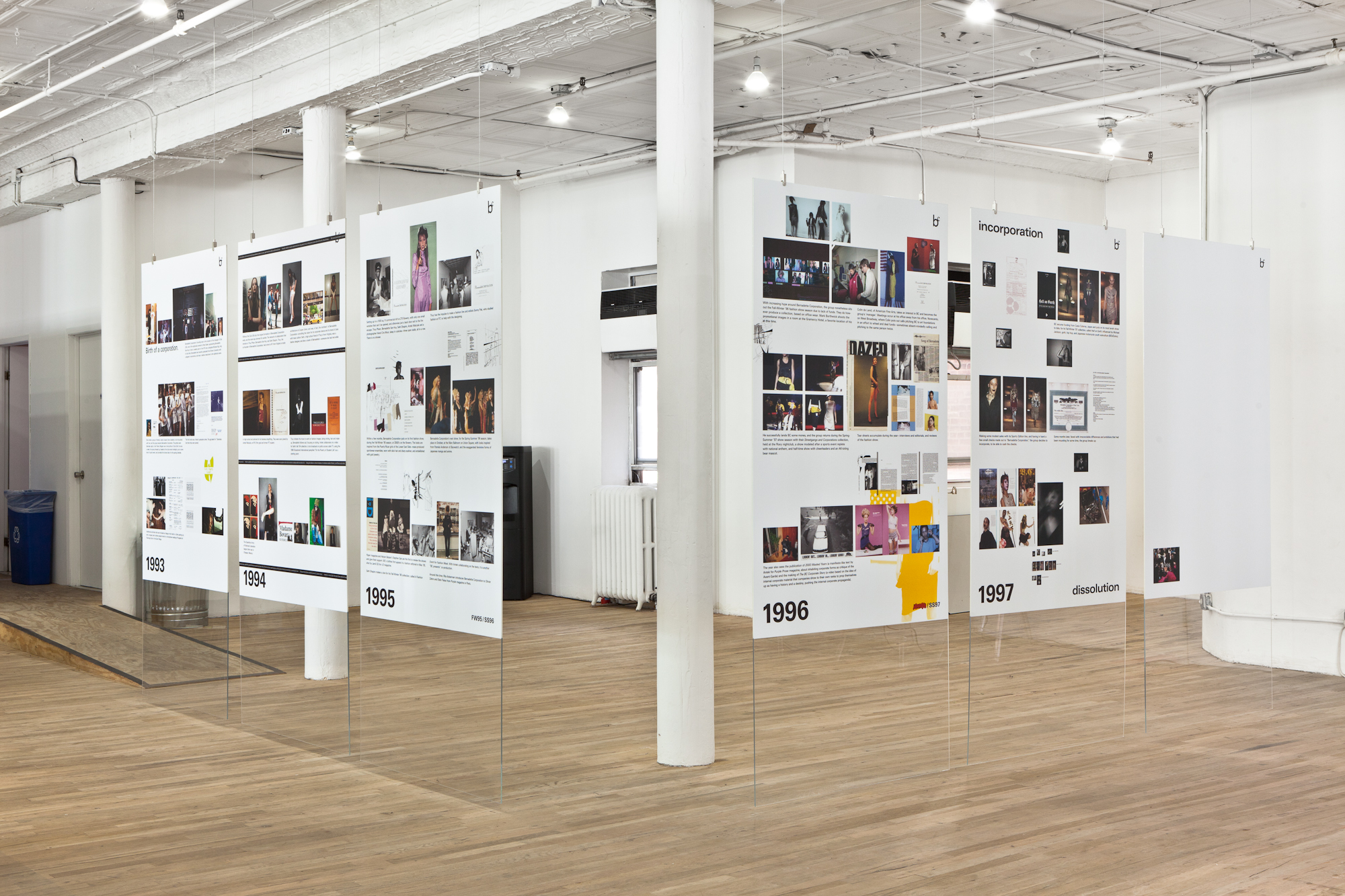 Six plexiglass slides holding large poster boards, arranged side-by-side such that they create two adjacent walls in the middle of a gallery space. The bottom left corner of each poster is marked with a year ranging from 1993-1997 and is filled with a collection of images and short texts. The rightmost poster is blank, save for a single photo.