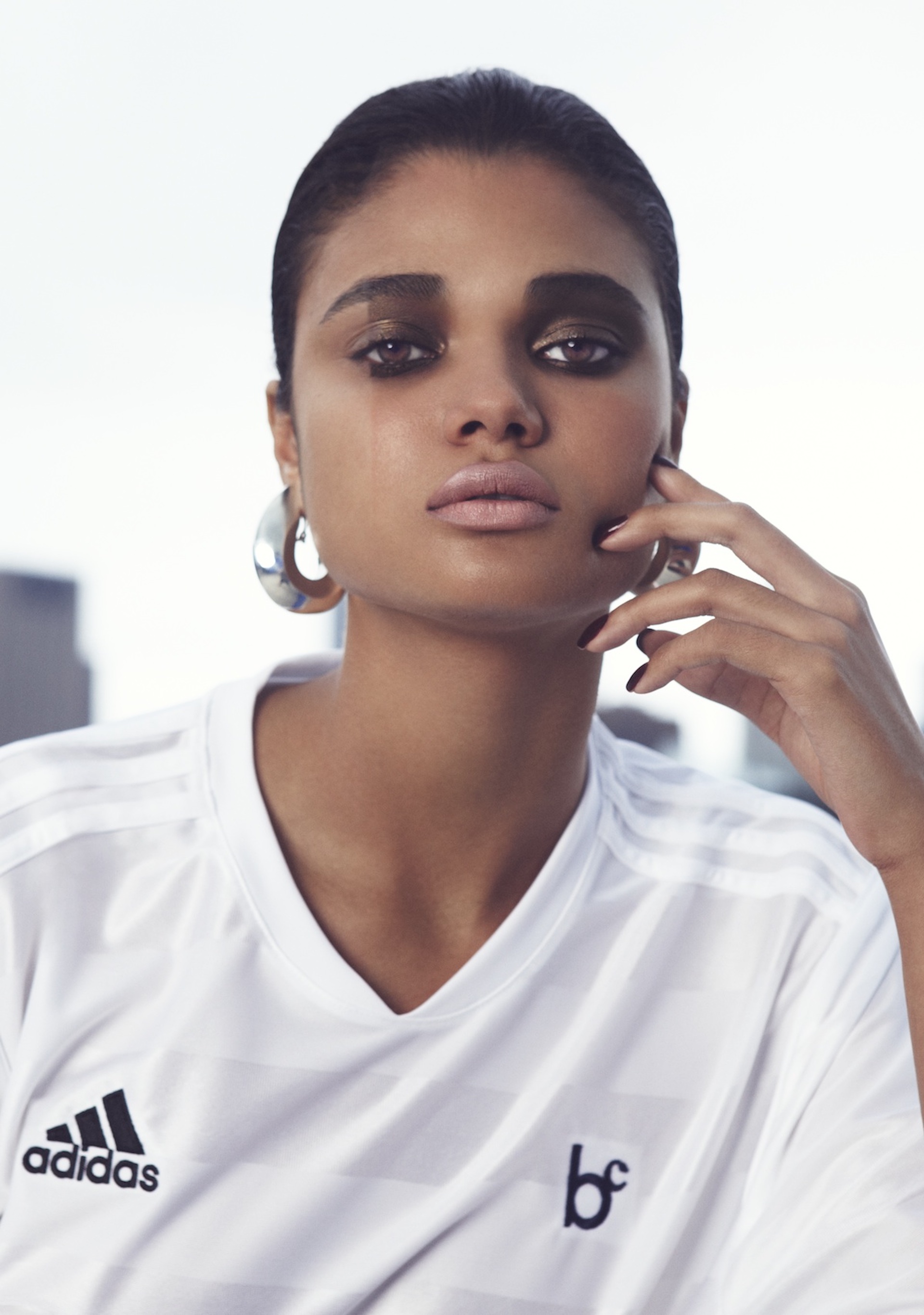 A photograph of a woman looking into the camera with one hand raised to her cheek. She wears a striped white Adidas soccer jersey with the 