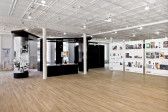 Three plexiglass and poster board timelines and a large black structure are visible within a gallery space. The timelines depict photos and texts arranged by year, from 1999-2001. The structure has an image of an office wrapped around one column and a TV is visible through the structure.