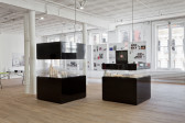 Two black display cases in the middle of a gallery space, with clear horizontal glass cut-outs, one taller than the other. Each display case houses a collection of mugs and other ephemera.