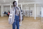 A female mannequin stands in a gallery space. It is wearing blue trousers and a grey fur coat over a blue checkered shirt. The shirt and jacket are left unbuttoned, exposing the mannequin's breasts, obscured by a large bone necklace.